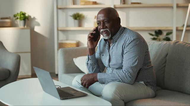 An elderly African American man sits comfortably on a couch, talking on the phone while working on a laptop. He appears focused and relaxed in a cozy living room setting.