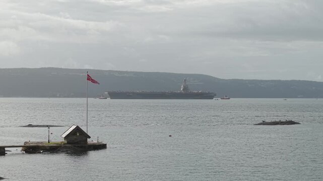 Island With A Flagpole And Hut With USS Gerald R. Ford Aircraft Carrier Cruising In The Background In Oslo, Norway. - wide shot
