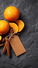 Three vibrant oranges, cinnamon sticks, and a blank tag are artistically arranged on a textured dark grey stone surface in this still life image.