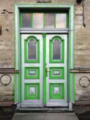 Double door with arched windows and green-gray paneling, set in a weathered stone wall with metal rings on each side, evoking vintage or historical architecture.