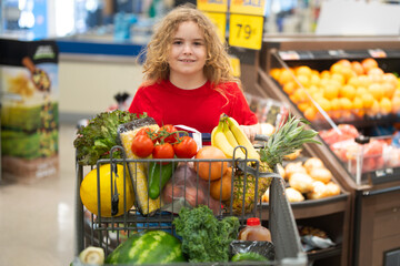 Child shopping with cart in grocery. Kid choosing fresh vegetables in grocery store. Child with food shopping in grocery. Kid selecting healthy produce at local shop. Child with fresh food in grocery.