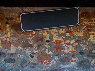 Empty black signboard on an old stone brick wall with rough texture, ready for text or logo placement.