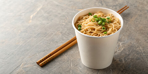 Instant noodle cup with chopstick and green onion garnish on textured surface, warm and appetizing meal