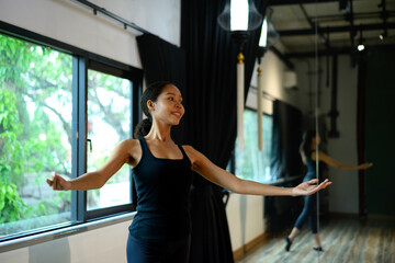 Graceful ballerina practicing dance pose in studio with arms extended elegantly near large window