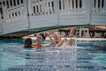 Joyful group of women and children splashing and laughing in a clear blue resort pool under a white bridge during a sunny summer day.