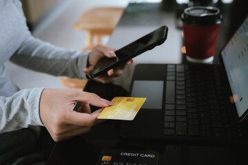 A young woman holding a credit card and coffee cup shops online using a laptop at a modern café, looking focused and happy.