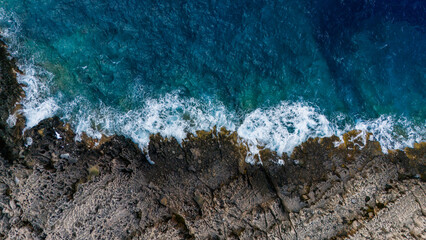 Aerial view of turquoise sea waves breaking on rugged rocky shoreline with white foam contrasting...