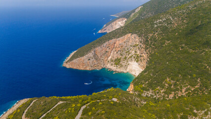 Overhead view of rugged coastline with steep cliffs turquoise cove deep blue sea and winding mountain road surrounded by lush green hills