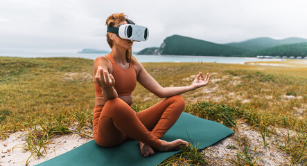 Woman meditating in lotus pose with VR headset, hands in mudra, sitting on yoga mat by sea. Concept of virtual wellness, mindfulness and digital meditation.