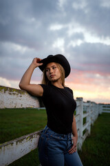 A young woman rests against a weathered fence while wearing a cowboy hat and casual attire during a vibrant sunset in the countryside. Generative AI.