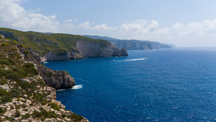Aerial view of rugged coastline with steep limestone cliffs green forest slopes turquoise coves and deep blue Ionian Sea under cloudy summer sky