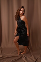 Full length portrait of pretty young barefoot brunette woman in black dress on brown background