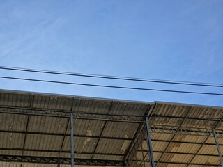 Low-angle outdoor view of a corrugated metal roof with exposed steel trusses of an industrial shed, set against a clear blue sky with two power lines crossing the top.