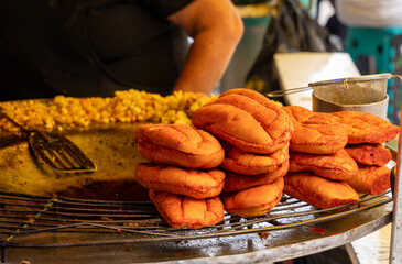 A person is standing behind a grill, cooking a pile of food that looks like bread or pastries. Historic center of Queretaro City, decorations and traditions to celebrate Mexico's Independence Day, col
