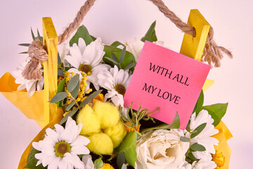 A vibrant floral arrangement features daisies, roses, and greenery in a yellow bag. A pink note attached expresses a warm sentiment, showcasing affection and love