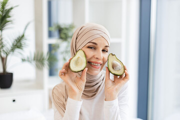 Smiling young Muslim woman holding avocado halves, wearing hijab in modern bright interior setting