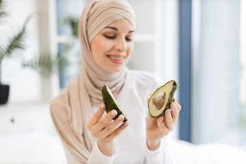 Smiling Muslim woman in beige hijab holding avocado halves indoors near natural lighting. Model adult woman appears cheerful and enjoys healthy lifestyle. Concept wellness, beauty, freshness, healthy.