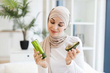 Young Muslim woman wearing headscarf holding avocado and lotion while smiling indoors. Promotes skincare, wellness, and natural product use for beauty routines.