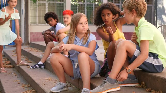 Portrait of cheerful preteen girls and boys having friendly conversation during gathering outdoors, sitting on steps on sunny summer day