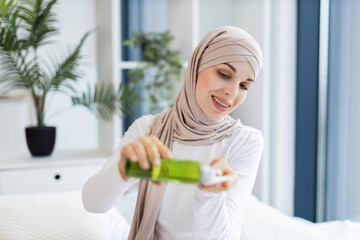 Young Muslim woman wearing hijab applying lotion for skincare in bright modern room. Focusing on self-care beauty and healthy lifestyle. Vibrant natural light and greenery create peaceful atmosphere.