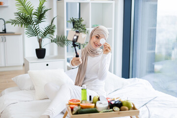 Muslim woman in loungewear using cotton pads and skincare products while recording self-care blog...