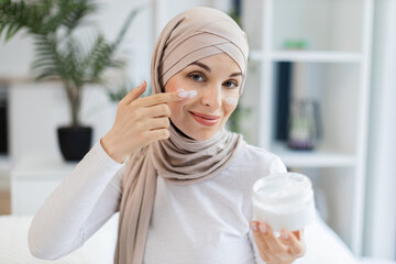 Smiling adult Muslim woman wearing hijab applying moisturizer cream on facial skin indoors. Promotes skincare, self-care, beauty regime, and wellness.