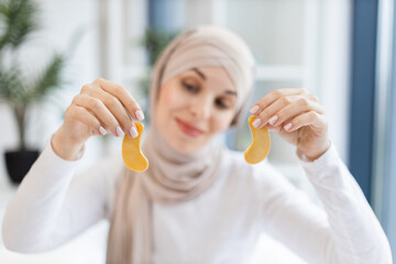 Fototapeta premium Young Muslim woman wearing hijab holding skincare gold eye patches. Bright indoor setting with focus on self-care routine. Promoting relaxation, beauty treatment.
