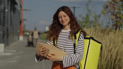 Smiling delivery woman with a thermal backpack carrying a cardboard box. Professional female courier walking along a modern city street on a sunny day providing a fast shipping service