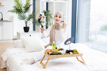 Young Muslim woman in her twenties wearing hijab records beauty vlog applying facial mask. Scene...