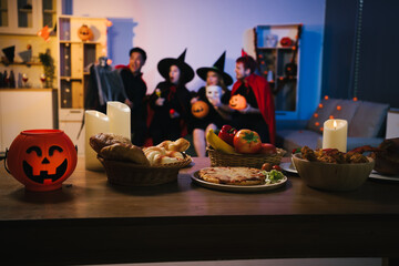 A cheerful group of friends dressed as witches and devils celebrate Halloween together,raising glasses of wine around a festive table with pumpkins,candles, bread,decorations in  cozy indoor setting
