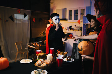 Four friends dressed as witches and devils celebrate Halloween together, holding pumpkins and decorations around a table with food and drinks. laugh and enjoy the spooky, festive atmosphere of party