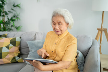 Asian senior woman using digital tablet in cozy living room at home.