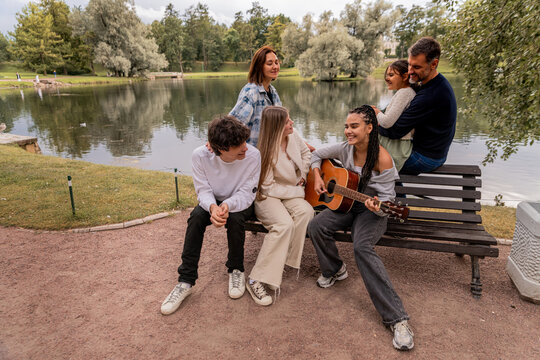 multigenerational group of friends plays guitar and sings by the lake.