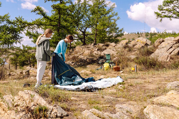 Fototapeta premium Two people setting up a tent in a rocky outdoor area surrounded by trees on a sunny day