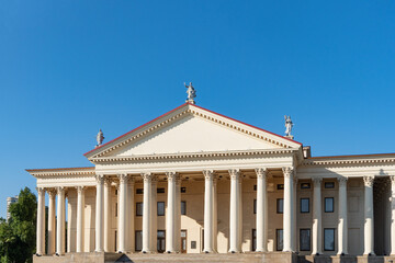 Fototapeta premium Neoclassical winter theatre building features impressive portico with tall Corinthian columns, pediment decorated with statues and red tile roof against clear blue sky. Sochi, Russia - June 28, 2025