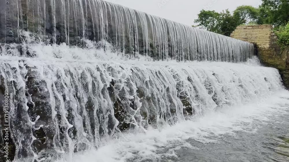 Poster waterfall pouring evenly over a stone wall, creating white frothy streams and splashes against dark 