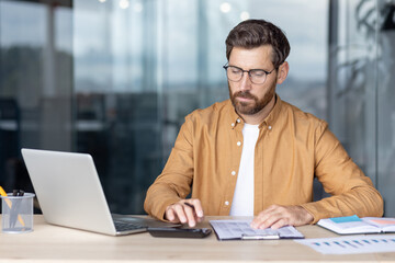 Fototapeta premium Focused businessman in eyeglasses sitting at an office desk, calculating financial data using a calculator and reviewing documents, managing personal finances or business expenses