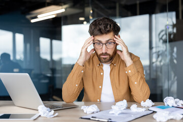 Young man having a headache at his desk, feeling frustrated and stressed, struggling with writer's block and a looming deadline, surrounded by crumpled papers