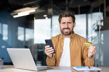 Young man at office desk holds smartphone and credit card, frowning in frustration over a failed online payment or banking error, stressed and worried about finances