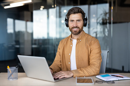 Young smiling business man with beard wearing headphones, typing on a laptop, enjoying remote work or online meeting at a desk in a modern corporate office