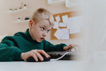 A boy with hearing aids focuses intently while playing a game on his laptop, showcasing modern...