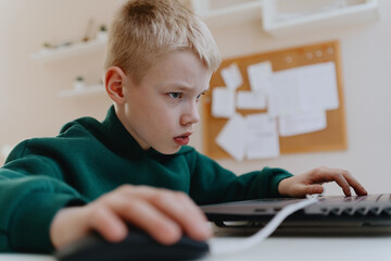 A boy with hearing aids focuses intently while playing a game on his laptop, showcasing modern...