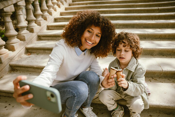Mother and son are holding ice cream and sitting on a blanket on the stairs while she is smiling and looking at the phone she is holding