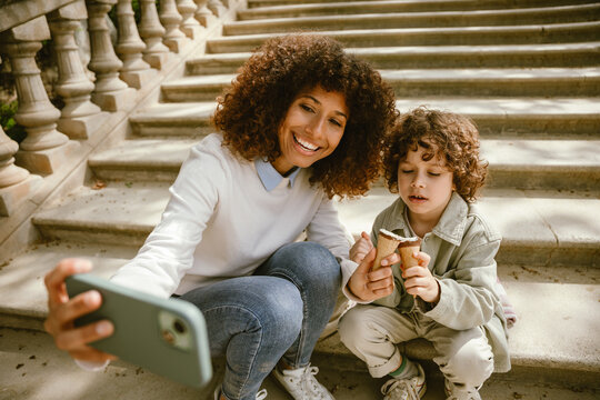 Mother and son are holding ice cream and sitting on a blanket on the stairs while she is smiling and looking at the phone she is holding