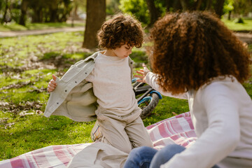 The son takes off his jacket while sitting on a blanket next to his mother, who reaches out to him