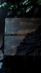 A moody, dark scene featuring a rustic wooden table with black sheer fabric or lace draped across the surface. Shadowy green foliage frames the backdrop