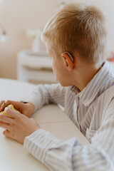 A boy with hearing aids in a button-down shirt enjoys a crisp apple at the kitchen counter. A natural moment of healthy snacking and inclusive childhood authenticity
