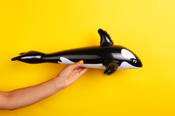 Hand with yellow nails holding a black and white inflatable whale toy against a bright yellow background, perfect for summer and playful themes.