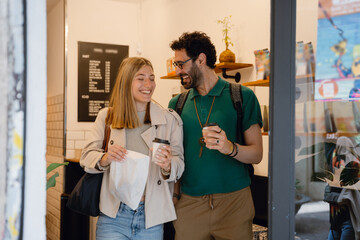 A man and a woman are holding cups and a bag while walking and laughing