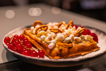 A delicious waffle topped with popcorn, marshmallows, and nuts, served with fresh red berries on a black plate.